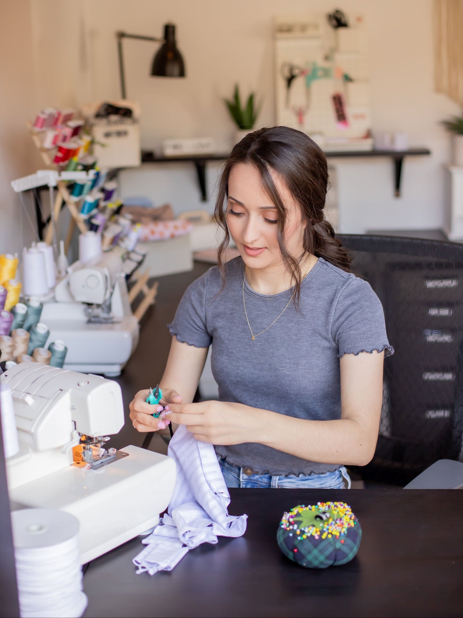 A woman in a gray t-shirt sits at a sewing table in a craft room, sewing a piece of white fabric. A white sewing machine is on the table next to her, with a colorful pin cushion on the desk in front of her.